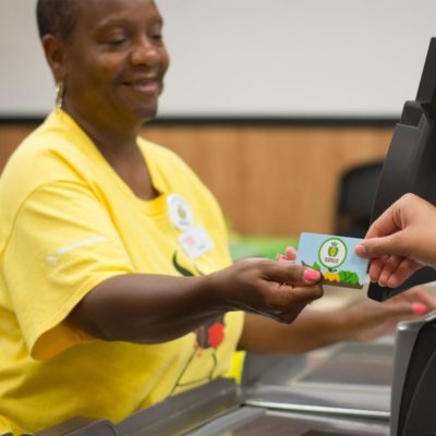 A shopper handing their Double Up Food Bucks card to a grocery store cashier in a yellow shirt with a Double Up Food Bucks badge on it.